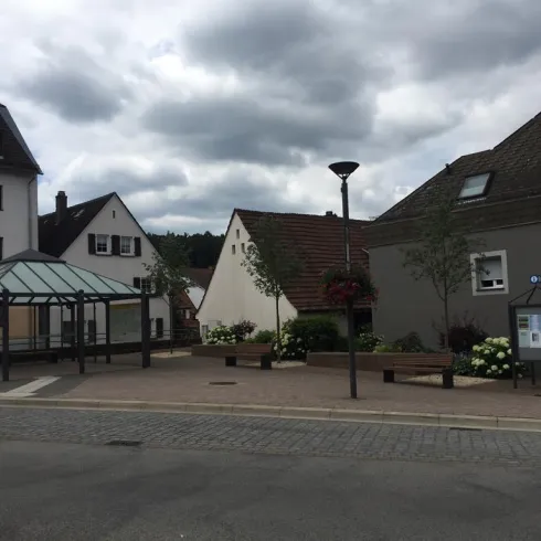 Eine kleine Stadtansicht mit modernen und traditionellen Gebäuden. Es gibt einen Wartebereich für Busse und einige Pflanzen in Blumentöpfen.