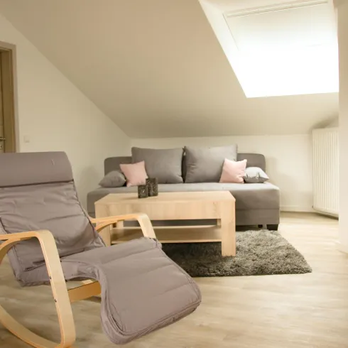 Ein gemütliches Wohnzimmer mit einer grauen Couch und einem hölzernen Tisch. Ein Holzstuhl lädt zum Entspannen ein, während viel natürliches Licht durch das Fenster fällt.