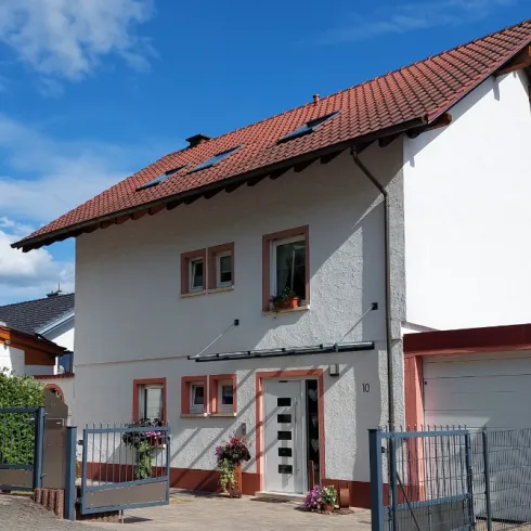 Ein modernes Haus mit einer roten Dachdeckung und weißen Wänden. Vor dem Haus gibt es einen kleinen Vorgarten mit Blumen und einem Zaun.