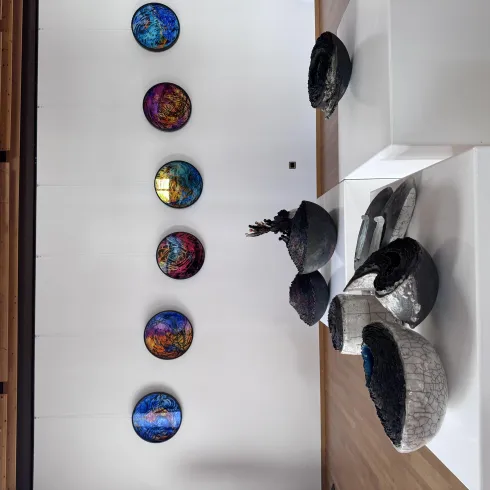 Zwei Männer stehen in einer Galerie und betrachten Kunstwerke. Um sie herum hängen weiße Skulpturen und weitere Objekte.