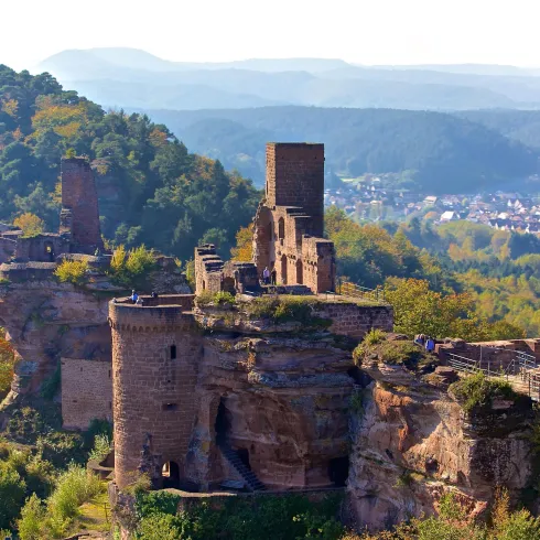 Eine alte Burg auf einem Hügel, umgeben von grünen Bäumen und sanften Bergen. Im Hintergrund sind weitere Landschaften zu sehen.