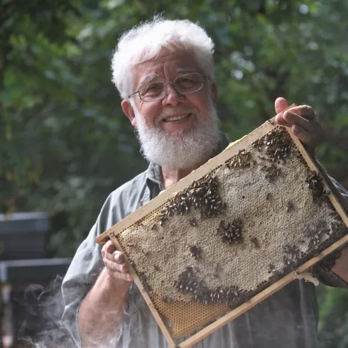Ein älterer Mann mit einem weißen Bart hält einen Wabenrahmen voller Honig. Im Hintergrund sind grüne Bäume zu sehen.