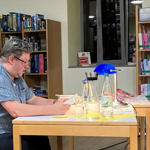 Ein Mann sitzt an einem Tisch in einer Bibliothek und liest etwas. Auf dem Tisch stehen Wasserflaschen und eine Lampe.