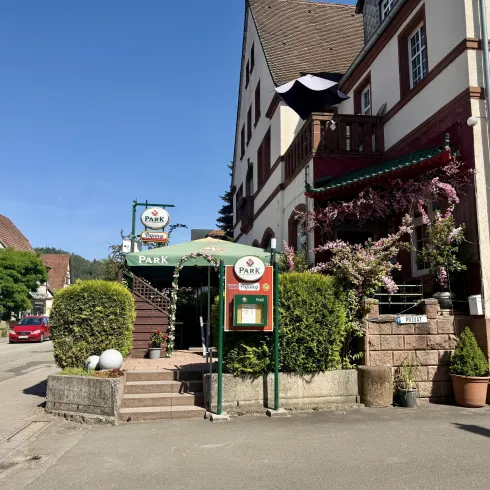Ein einladendes Restaurant mit einer schönen Terrasse und grünen Pflanzen. Die Fassade ist hell, und der Himmel ist blau.