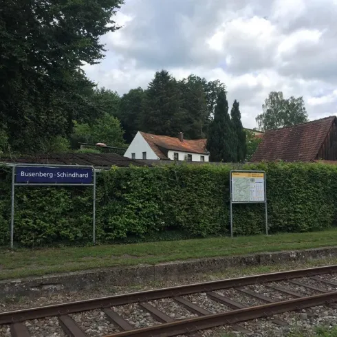Ein Bahnhofsschild für Busenberg-Schindhard ist sichtbar, umgeben von Büschen und Bäumen. Im Hintergrund sind einige Häuser zu sehen, und der Himmel ist bewölkt.