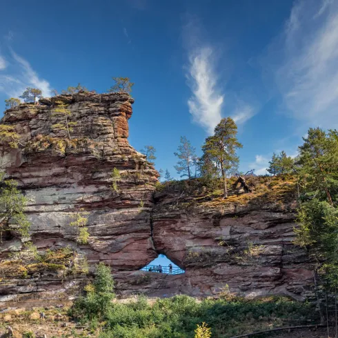 Ein beeindruckender Felsformation mit einem natürlichen Fenster. Umgeben von Bäumen und einem klaren Himmel.