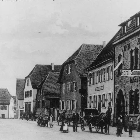 Eine historische Straßenszene mit mehreren Gebäuden. Menschen und Pferdewagen sind ebenfalls zu sehen.