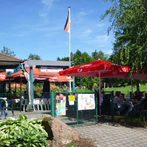 Ein gemütliches Café mit roten Sonnenschirmen und einer deutschen Flagge. Viele Gäste genießen die Sonne und die entspannte Atmosphäre im Freien.