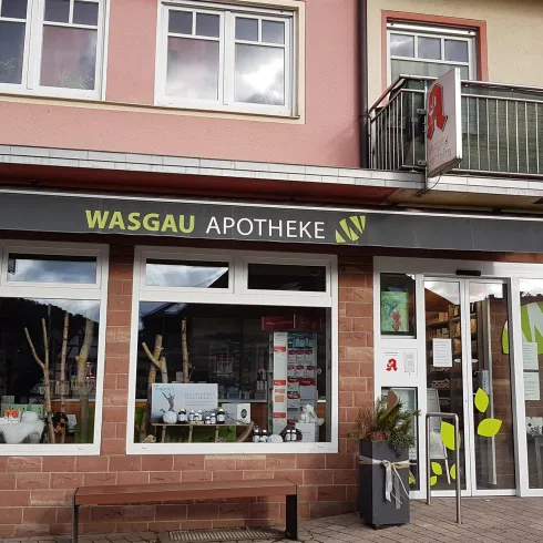 Eine Apotheke mit der Aufschrift "WASGAU APOTHEKE". Der Eingang ist klar sichtbar, und im Fenster sind verschiedene Produkte ausgestellt.