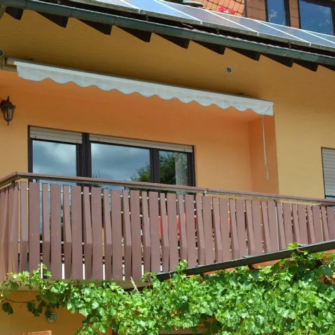 Ein sonniger Balkon mit Holzgeländer und einer weißen Markise. Grünes Blattwerk umrahmt den Bereich.