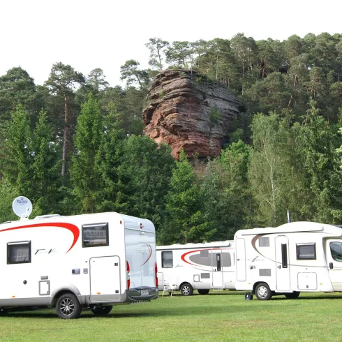 Ein Campingplatz mit mehreren Wohnmobilen auf grünem Rasen. Im Hintergrund ragt ein großer Felsen zwischen Bäumen empor.