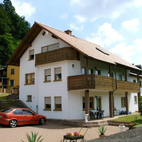 Ein modernes Haus mit Holzverzierungen und einem großen Balkon. Vor dem Haus steht ein rotes Auto auf einem gepflasterten Parkplatz.