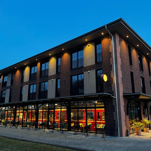 Ein modernes Gebäude mit roten Ziegelfassaden und großen Fenstern. Die Fassade ist gut beleuchtet und ein Zug ist sichtbar.