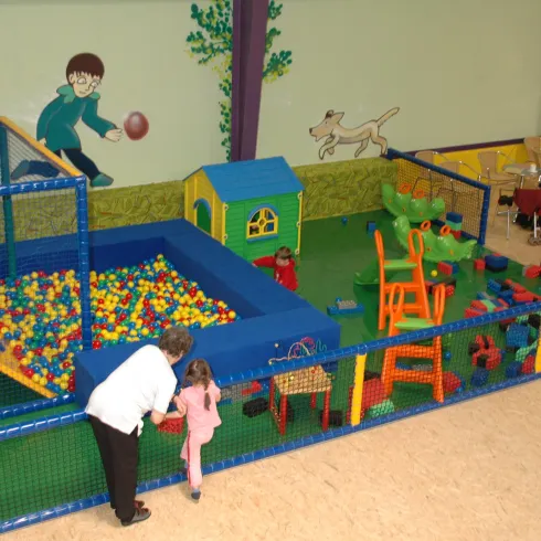 Ein bunter Spielplatz mit einem Bällebad und Rutschen für Kinder. Es gibt Spielgeräte und eine kleine Häuschen, die den Raum lebendig machen.
