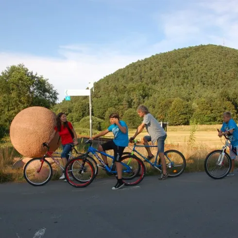Eine Gruppe von Kindern und Jugendlichen steht auf Fahrrädern an einem Kreuzungspunkt. Im Hintergrund ist ein grüner Hügel und eine große, runde Skulptur zu sehen.