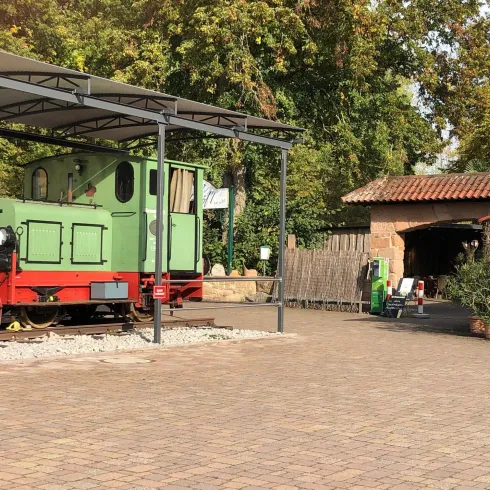 Eine grüne Lokomotive steht unter einem Vordach auf einem gepflasterten Platz. Im Hintergrund ist ein rustikales Gebäude und grüne Bäume zu sehen.