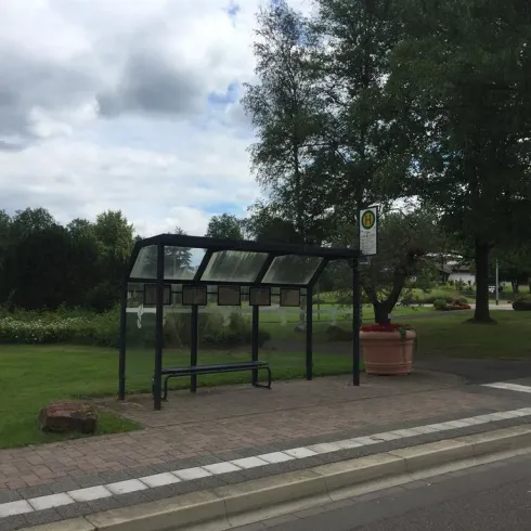 Eine Bushaltestelle in einer grünen Umgebung mit Bäumen. Über der Bank steht ein Dach, und es gibt eine Informationssäule daneben.