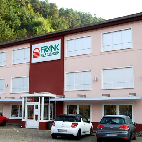 Ein modernes Geschäftshaus mit dem Namen „FRANK Korbwaren“. Vor dem Gebäude stehen mehrere Autos auf einem Parkplatz.