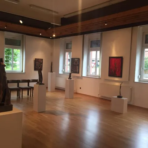 Ein heller Ausstellungsraum mit Skulpturen auf Podesten und Bildern an den Wänden. Große Fenster lassen viel Tageslicht herein.