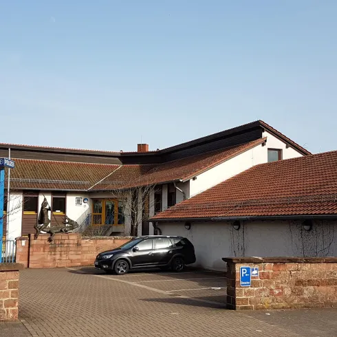 Ein Gebäude mit aus rotem Stein und einem flachen Dach. Ein schwarzes Auto steht auf dem Parkplatz vor dem Eingang.