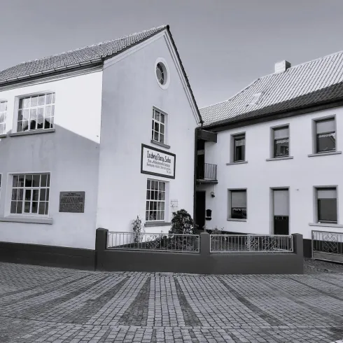 Eine Szene mit zwei weißen Gebäuden, die nebeneinander stehen. Der Platz dazwischen ist gepflastert und bietet einen klaren Blick auf die Architektur.