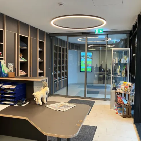 Ein moderner Empfangsbereich mit einem Schreibtisch, Regalen und einer Glaswand. Im Hintergrund ist ein Bildschirm sichtbar, sowie ein kleiner Bereich mit Zeitschriften und Snacks.