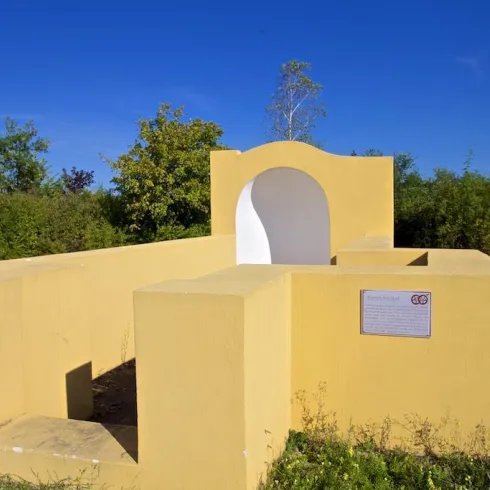 Eine moderne, gelbe Architekturstruktur mit einem offenen Bogen. Umgeben von grüner Vegetation und blauem Himmel.