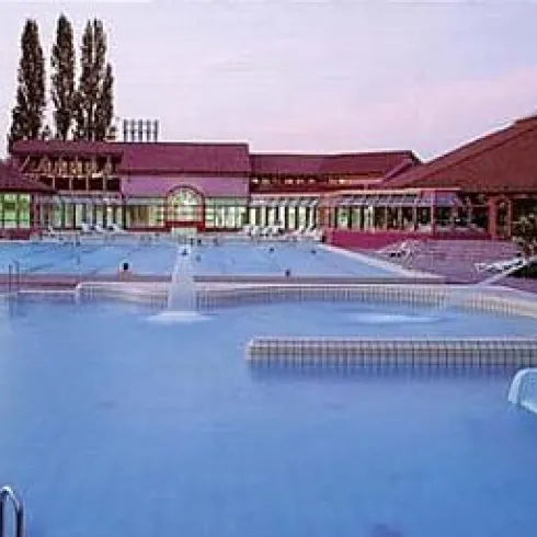 Ein Schwimmbad mit mehreren Rutschen und einem klaren Wasser. Im Hintergrund ist ein großes Gebäude mit Fenstern und Bäumen zu sehen.