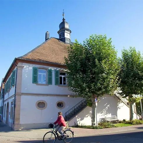 Ein historisches Gebäude mit einem markanten Turm und grünen Fensterläden. Im Vordergrund fährt ein Radfahrer vorbei, umgeben von Bäumen und gutem Wetter.