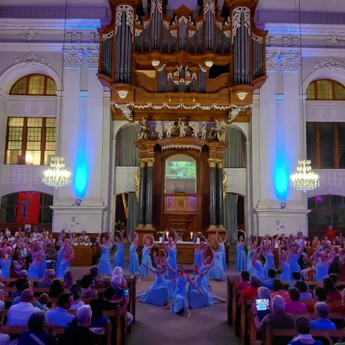 Eine festliche Aufführung in einer prächtigen Kirche mit einem großen Orgelretabel. Tänzerinnen in blauen Kleidern führen vor einem begeisterten Publikum auf.