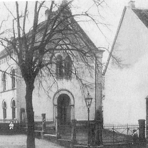 Ein historisches Gebäude mit einer markanten Fassade und großen Fenstern. Im Vordergrund sind Bäume und eine Straße zu sehen.