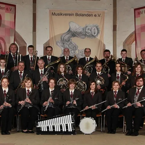 Eine große Musikgruppe steht in formellen Anzügen zusammen. Im Hintergrund sind dekorative Banner zu sehen.
