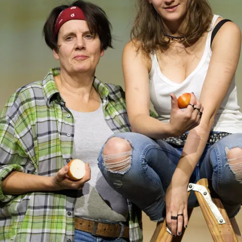 Zwei Frauen stehen zusammen, eine hält einen Apfel in der Hand. Die andere sitzt auf einer Leiter und zeigt ebenfalls einen Apfel.