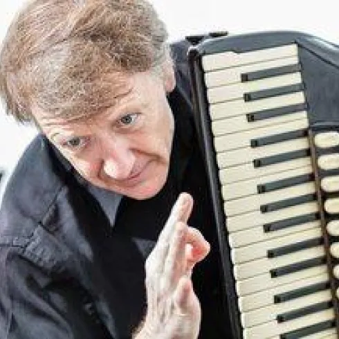 Ein Mann spielt Akkordeon und zeigt mit der Hand eine Geste. Er trägt ein schwarzes Hemd und hat lockiges Haar.