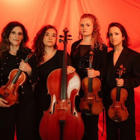 Vier Musikerinnen stehen vor einem roten Hintergrund und halten ihre Instrumente. Sie tragen elegante schwarze Kleidung und scheinen bereit für einen Auftritt zu sein.
