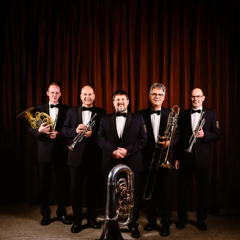 Eine Musikgruppe aus fünf Männern in eleganten Anzügen steht vor einem braunen Hintergrund. Sie halten verschiedene Blechblasinstrumente, darunter eine Tuba und eine Trompete.
