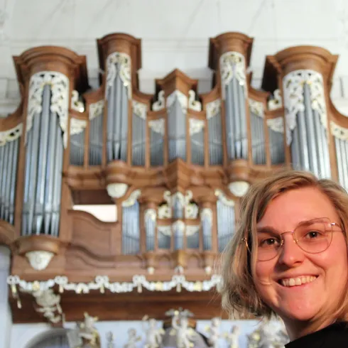 Eine Person lächelt vor einer großen Orgel. Die Orgel ist kunstvoll gestaltet und hat viele Pfeifen.
