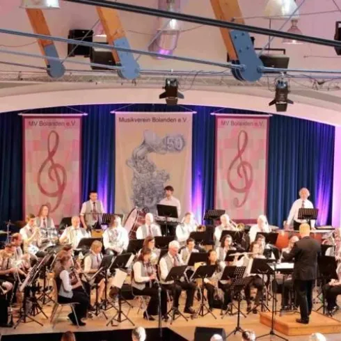 Ein Blasorchester spielt auf einer Bühne mit buntem Licht. Im Hintergrund sind Musikinstrumente und Notenblätter sichtbar.