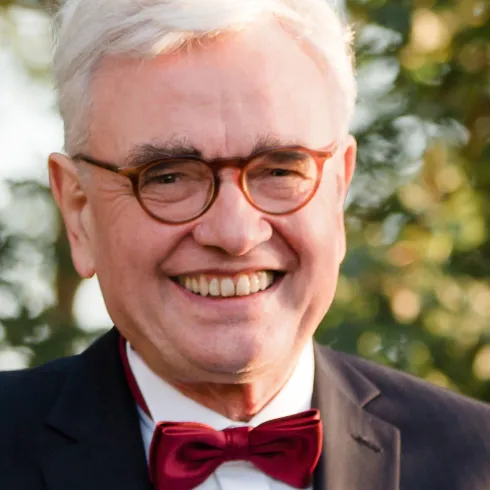 Ein lächelnder Mann mit grauen Haaren trägt einen schwarzen Anzug und eine rote Fliege. Im Hintergrund sind verschwommene grüne Bäume zu sehen.