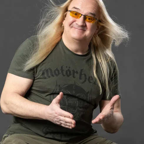 Ein lächelnder Mann mit langen, blonden Haaren sitzt auf einem Hocker. Er trägt eine grüne T-Shirt und auffällige gelbe Sonnenbrille.