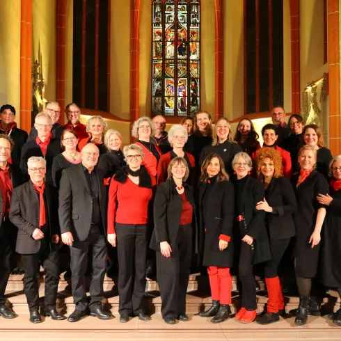 Eine Gruppe von Sängern steht auf einer Treppe in einer Kirche. Sie tragen schwarze Kleidung mit roten Akzenten und lächeln in die Kamera.