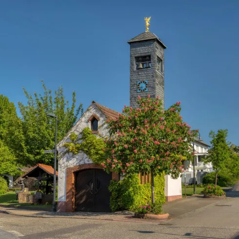 Ein charmantes Gebäude mit einem Uhrenturm und üppigem Grün umgeben. Der klare Himmel und die ruhige Straße verleihen der Szene eine friedliche Atmosphäre.