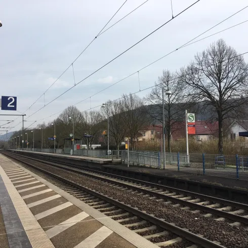 Ein Bahnhof mit einem Gleis und einer Wartebereich. Im Hintergrund sind Bäume und einige Gebäude zu sehen.