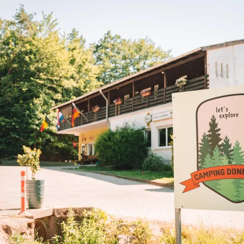 Ein Campingplatz mit einem großen Schild, das "Camping Donnersberg" zeigt. Im Hintergrund sieht man ein Gebäude, umgeben von Bäumen und Pflanzen.
