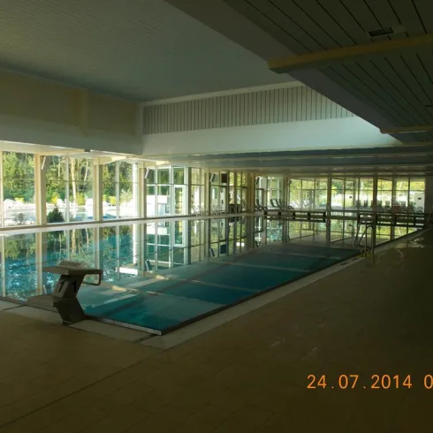 Ein helles Hallenbad mit großen Fenstern und einem klaren Wasserbecken. An der Seite befinden sich Sprungbretter und Liegen.