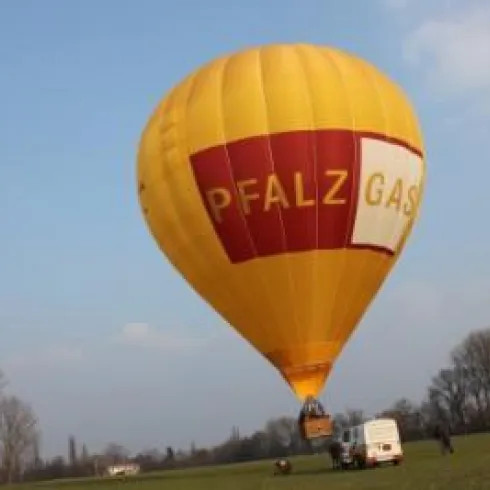 Ein gelber Heißluftballon mit dem Schriftzug „PFALZ GAS“ schwebt über einem grünen Feld. Im Hintergrund sind Bäume und ein blauer Himmel zu sehen.