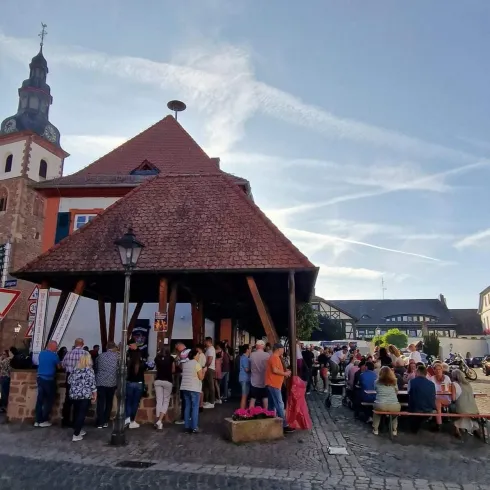 Ein belebter Marktplatz mit vielen Menschen und einem historischen Gebäude. Im Hintergrund ist ein erhöhter Kirchturm zu sehen.