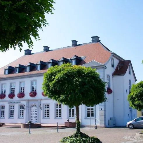 Ein historisches Gebäude mit einem roten Dach und vielen Fenstern. Vor dem Haus stehen runde Bäume und es ist sonnig.