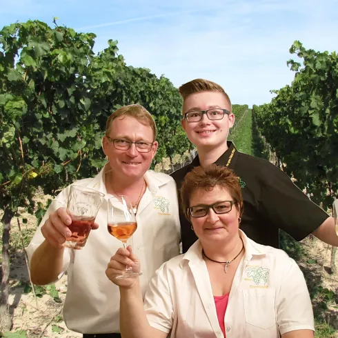 Eine fröhliche Familie steht in einem Weinberg und hält Gläser mit Wein in der Hand. Im Hintergrund sind grüne Weinreben und ein blauer Himmel zu sehen.