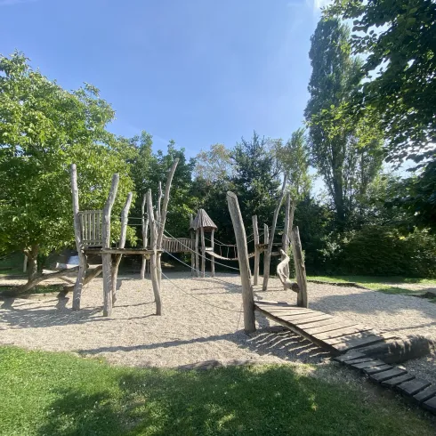 Ein Spielplatz mit Holzstrukturen und einer Sandfläche. Umgeben von Bäumen und einem klaren blauen Himmel.